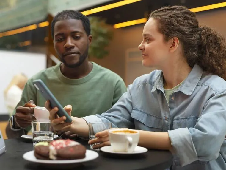 Pareja en un café revisando juntos el celular, con pasteles sobre la mesa